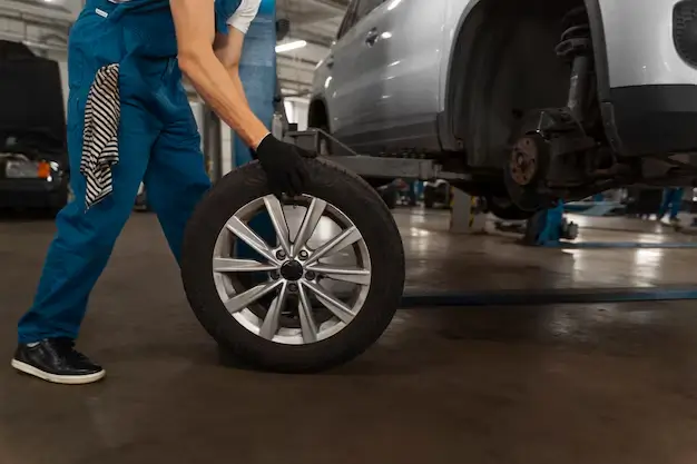 A mechanic in a workshop rolling a car tire towards a lifted vehicle for installation or replacement.