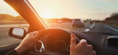 A driver's hands gripping the steering wheel while driving on a highway at sunset, with cars ahead and a warm golden glow illuminating the scene.